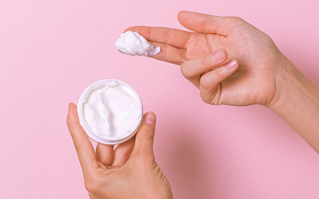pink background, a woman's hands holding lotion container in one hand and some lotion on the finger tips of the other hand